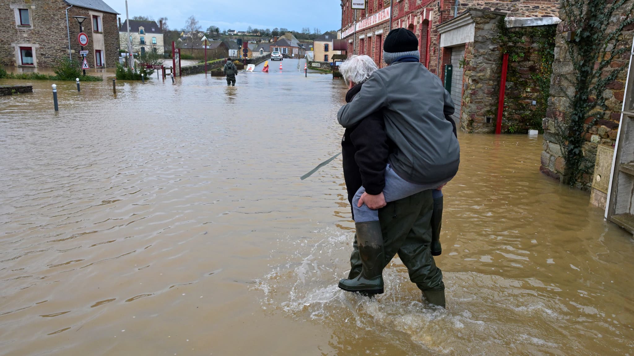 Un homme porte une personne sur son dos alors qu'ils traversent une route inondée suite au débordement de la Vilaine, à Pont-Rean, le 27 janvier 2025.
