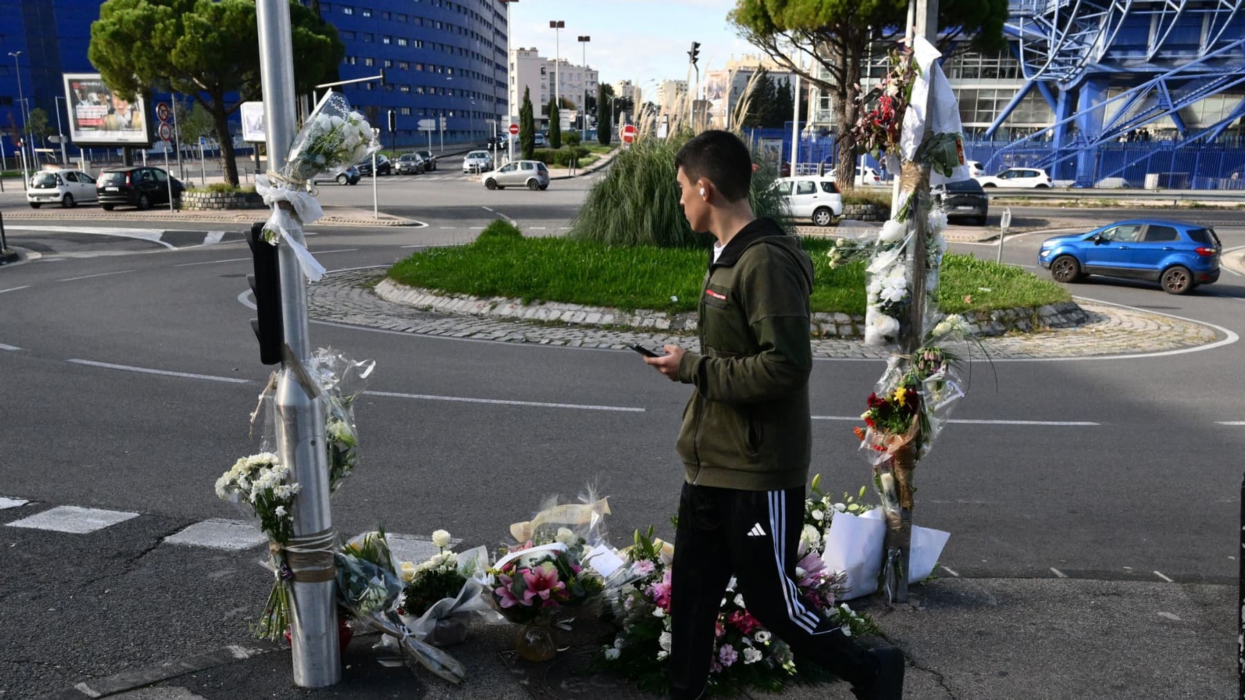 Un piéton passe devant des bouquets de fleurs déposés devant un mémorial de fortune où Mehdi Kessaci a été assassiné, au rond-point Claudie-Darcy, à Marseille, le jour de ses funérailles le 18 novembre 2025.