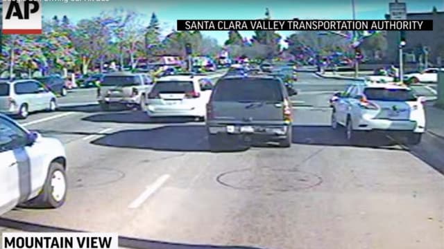 A droite sur l'image, la Google Car avance doucement, clignotant enclenché, pour changer de file, alors que le bus des transports publics continue de progresser sur sa file.