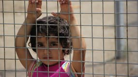 Une petite fille, les mains accrochées à la clôture regarde en direction des officiers du camp.
