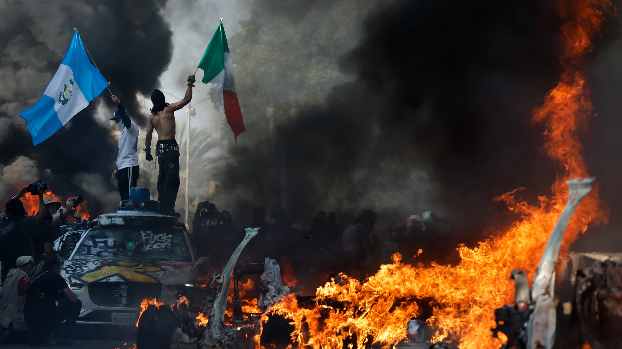 Des manifestants brandissent des drapeaux tandis que des voitures Waymo incendiées et vandalisées bordent la rue, le 8 juin 2025 à Los Angeles, en Californie. Des manifestants brandissent des drapeaux tandis que des voitures Waymo incendiées et vandalisées bordent la rue, le 8 juin 2025 à Los Angeles, en Californie.