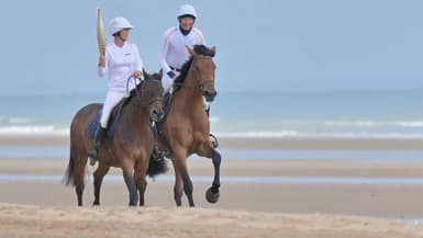 la cavalière Pénélope Leprévost (g) porte la flamme olympique, accompagnée de l'humoriste Nicolas Canteloup, sur la plage d'Omaha Beach, le 30 mai 2024 à Saint-Laurent-sur-Mer, dans le Calvados