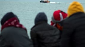 Des migrants assis sur la plage regardent un canot de sauvetage ramener des clandestins, secourus lors de la traversée de la Manche, sur une plage de Dungeness, sur la côte sud-est de l’Angleterre le 24 novembre 2021 