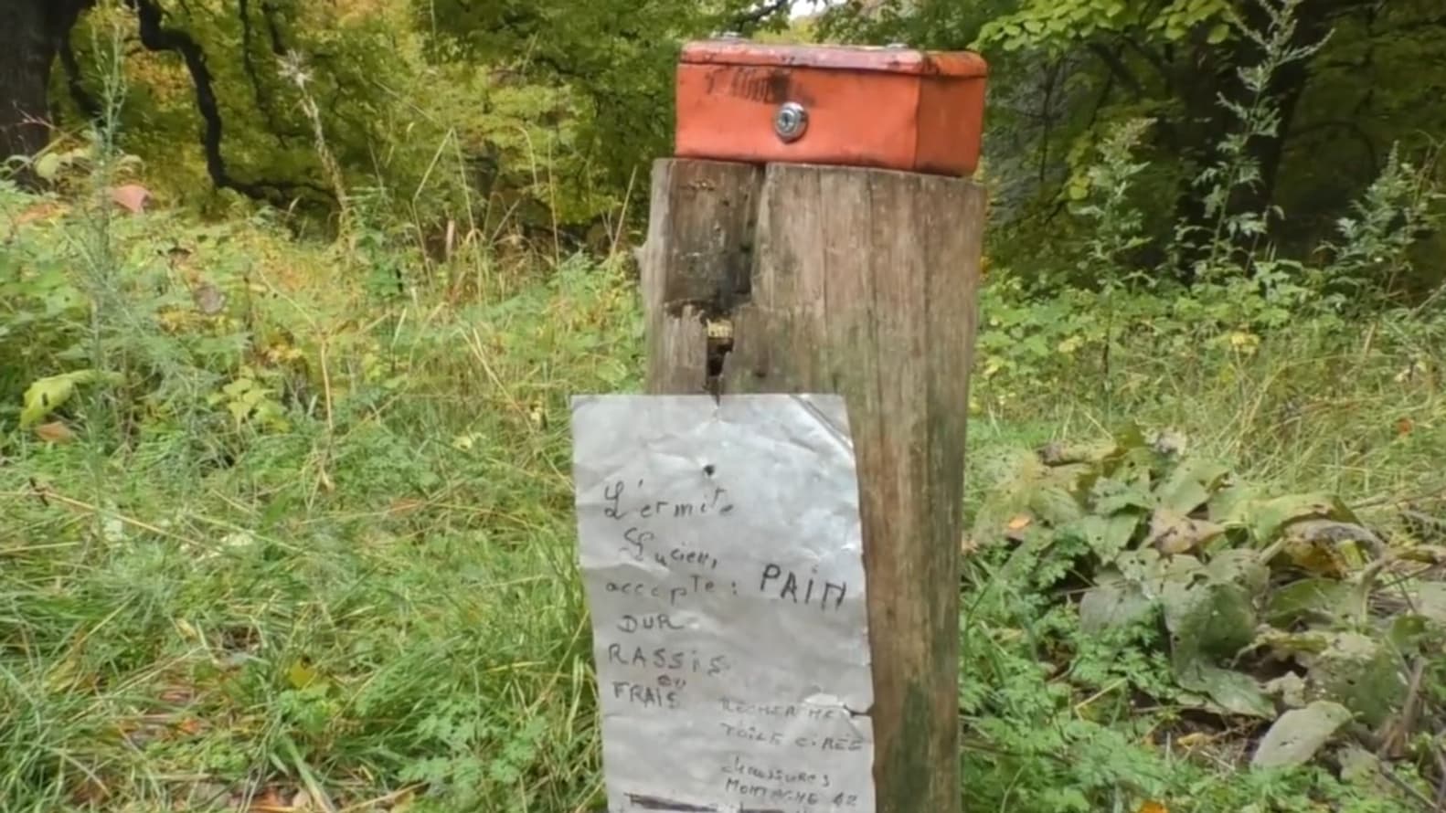 Devant la cabane de Lucien, sur un tronc, un écriteau où on peut lire "L'ermite Lucien accepte le pain dur rassis ou frais" à Saint-Etienne-les-Orgues".