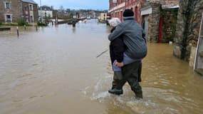 Un homme porte une personne sur son dos alors qu'ils traversent une route inondée suite au débordement de la Vilaine, à Pont-Rean, le 27 janvier 2025.