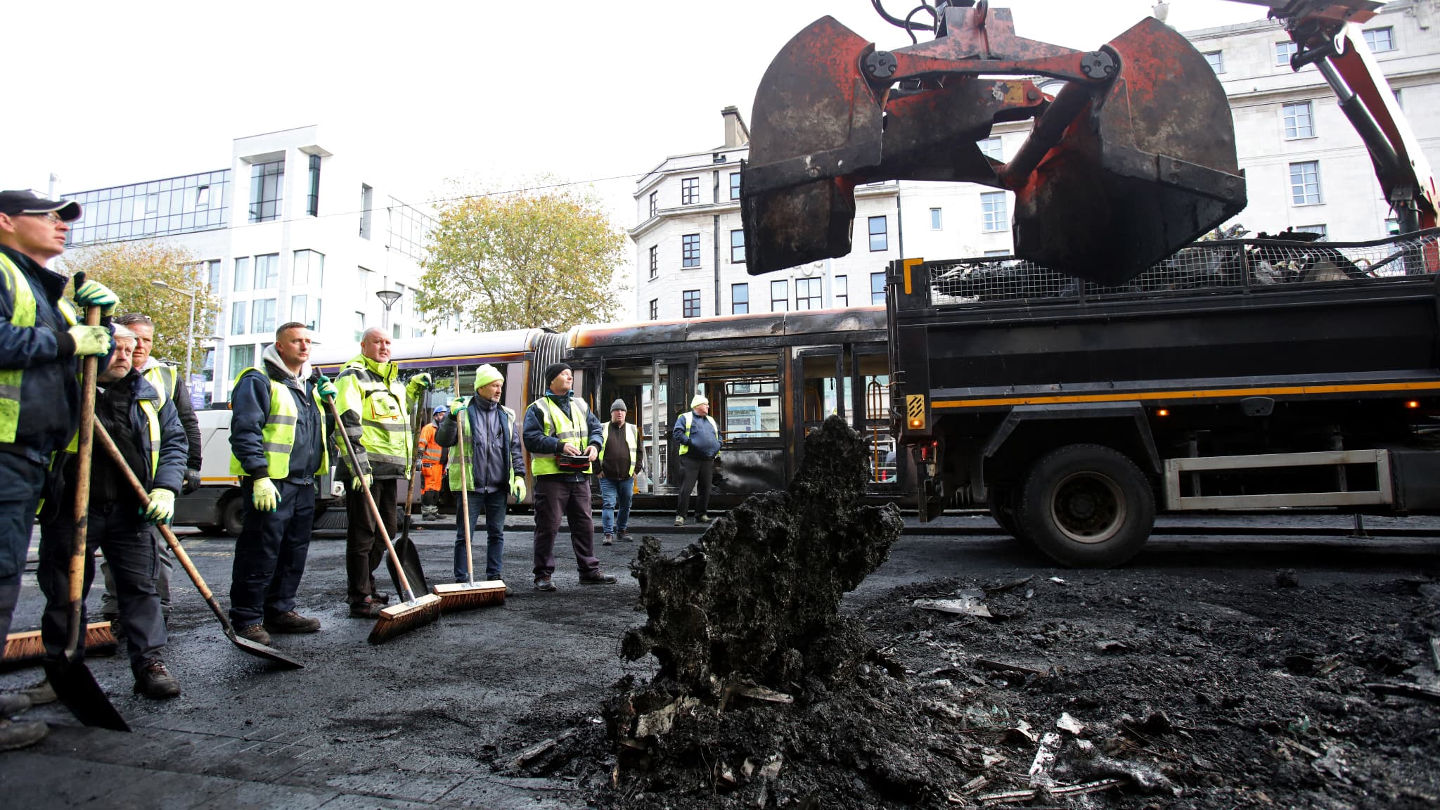 Des ouvriers enlèvent les restes d'un bus incendié d'une route alors qu'un tramway Luas endommagé par le feu se trouve à l'arrière-plan, sur la rue O'Connell à Dublin, le 24 novembre 2023, après une nuit de protestations. Des ouvriers enlèvent les restes d'un bus incendié d'une route alors qu'un tramway Luas endommagé par le feu se trouve à l'arrière-plan, sur la rue O'Connell à Dublin, le 24 novembre 2023, après une nuit de protestations.