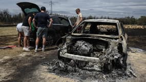 Des personnes se tiennent près d'une voiture calcinée dans une zone de loisirs touchée par un incendie de forêt à Sigean, le 27 juillet 2025. 