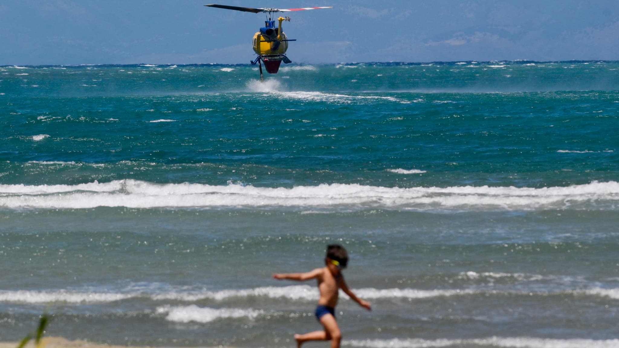 Un garçon joue sur le front de mer alors qu'un hélicoptère de lutte contre les incendies plane au-dessus de la mer pour remplir de l'eau afin de lutter contre les feux de forêt près du village de Loutses sur l'île grecque de Corfou, le 27 juillet 2023. Un garçon joue sur le front de mer alors qu'un hélicoptère de lutte contre les incendies plane au-dessus de la mer pour remplir de l'eau afin de lutter contre les feux de forêt près du village de Loutses sur l'île grecque de Corfou, le 27 juillet 2023.
