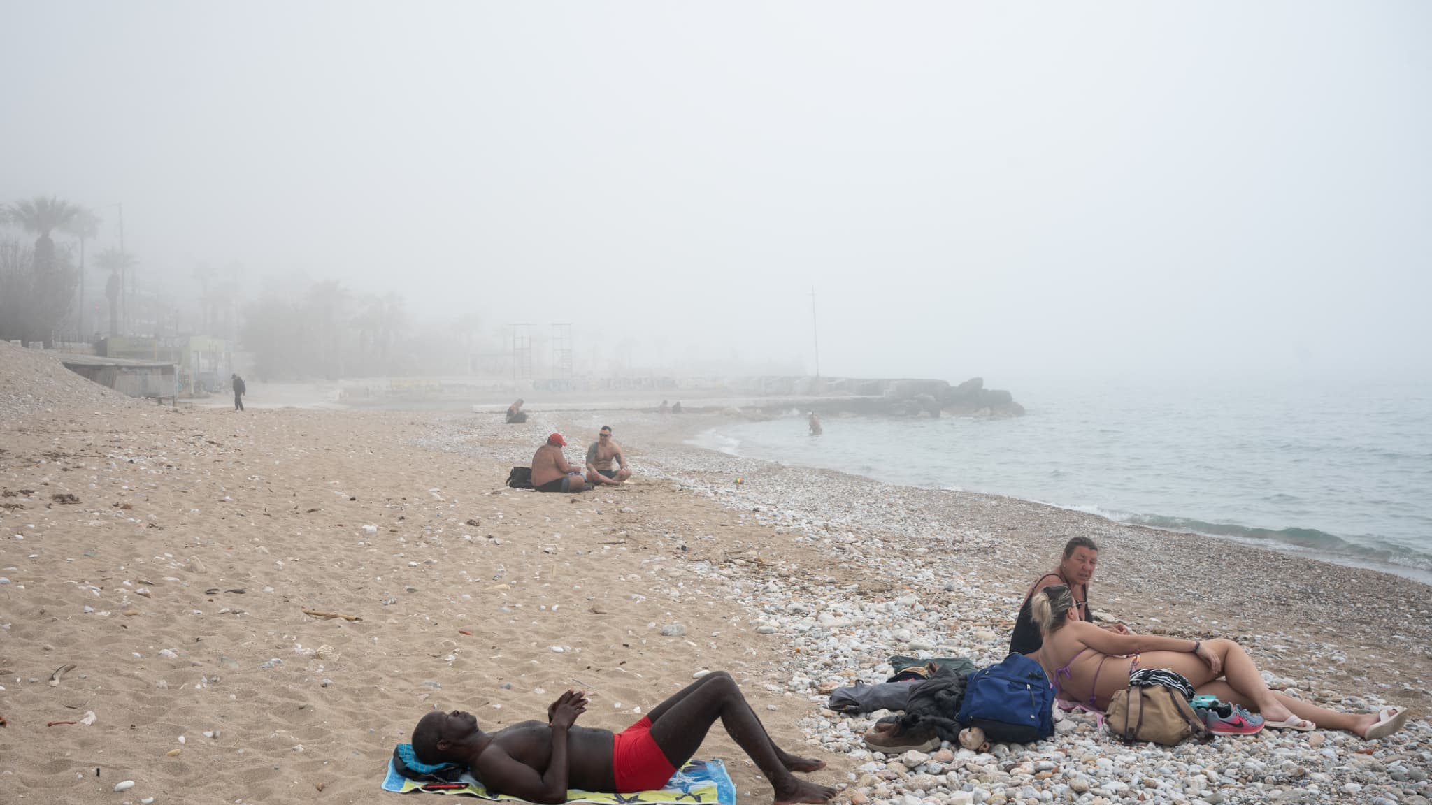 Des personnes sur une plage alors que la poussière et le brouillard couvrent l'horizon dans la banlieue de Palaio Faliro, en Grèce, au sud d'Athènes, le 15 mars 2025.