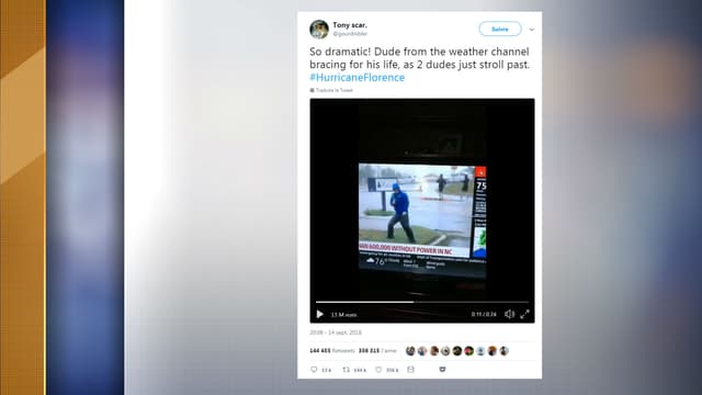 Un journaliste de The Weather Channel semble lutter contre le vent, alors que deux hommes marchent tranquillement derrière lui.