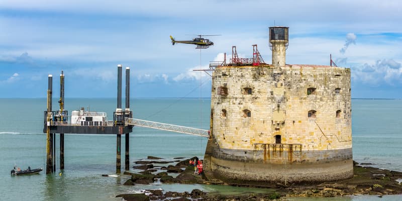 Depuis les airs, un hélicoptère transporte des bennes contenant du béton coulé en immergé, directement sous l’eau, destiné à combler une cavité creusée par les tempêtes au pied du fort Boyard (Charente-Maritime).