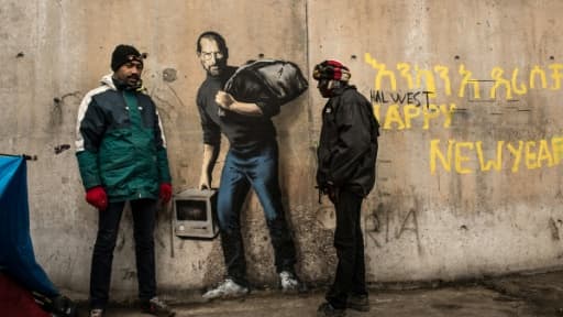 Des migrants devant une oeuvre de l'artiste britannique Bansky photographiée le 12 décembre 2015 et située à l'entrée de la "Jungle" de Calais, représente Steve Jobs (Apple), qui porte un baluchon et un vieil ordinateur