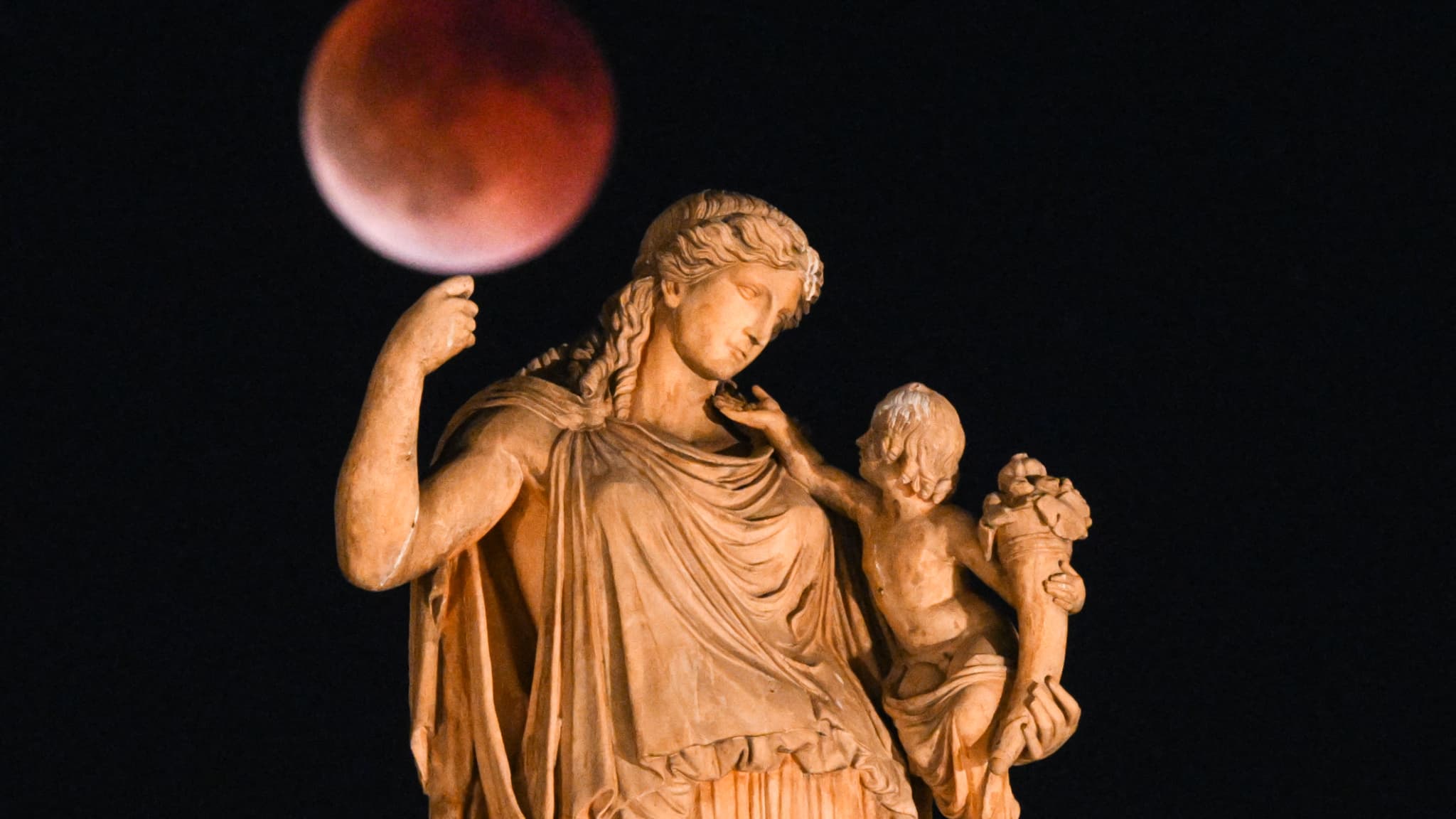 Cette photographie montre une pleine lune, également appelée "lune de sang", lors d'une éclipse lunaire, avec au premier plan une statue de la déesse grecque antique Irène tenant un enfant, dans le centre d'Athènes, le 7 septembre 2025. Cette photographie montre une pleine lune, également appelée "lune de sang", lors d'une éclipse lunaire, avec au premier plan une statue de la déesse grecque antique Irène tenant un enfant, dans le centre d'Athènes, le 7 septembre 2025.