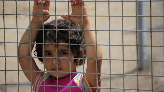 Une petite fille, les mains accrochées à la clôture regarde en direction des officiers du camp.