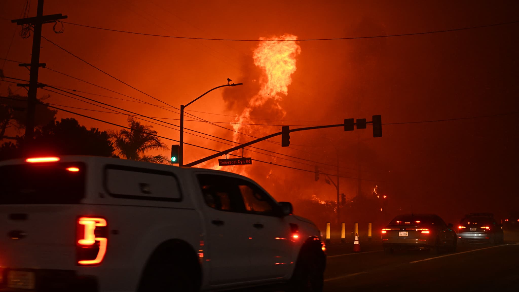 Des voitures passent devant le feu qui envahit l'intersection de Temescal Canyon et de Pacific Coast Highway lors de l'incendie de Palisades à Pacific Palisades, Californie, le 7 janvier 2025.