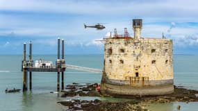 Depuis les airs, un hélicoptère transporte des bennes contenant du béton coulé en immergé, directement sous l’eau, destiné à combler une cavité creusée par les tempêtes au pied du fort Boyard (Charente-Maritime). 